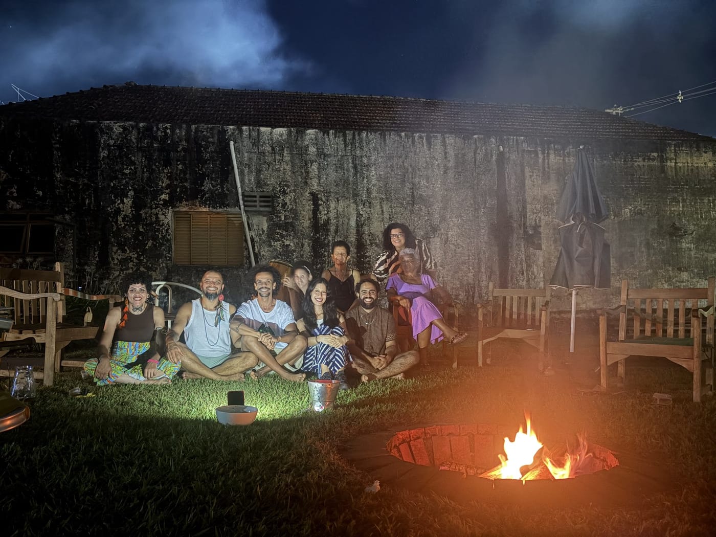 Grupo sentado na grama em frente à fogueira, na noite do Luau