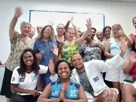 Grupo numeroso acenando em sala, junto de profissionais de saúde